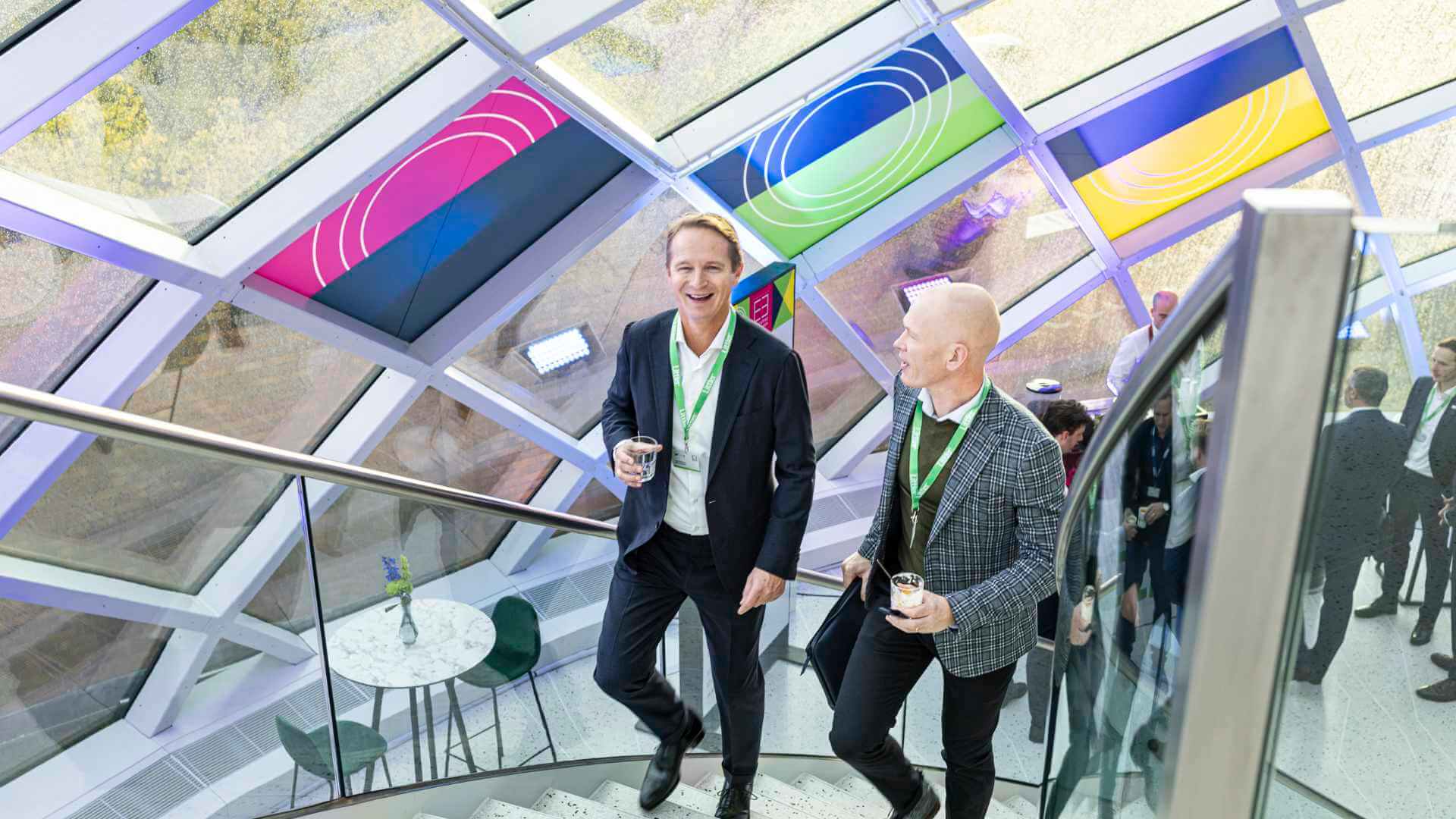 Two corporate event attendees networking and laughing on a staircase inside a contemporary conference venue with colorful geometric glass ceiling panels overhead.
