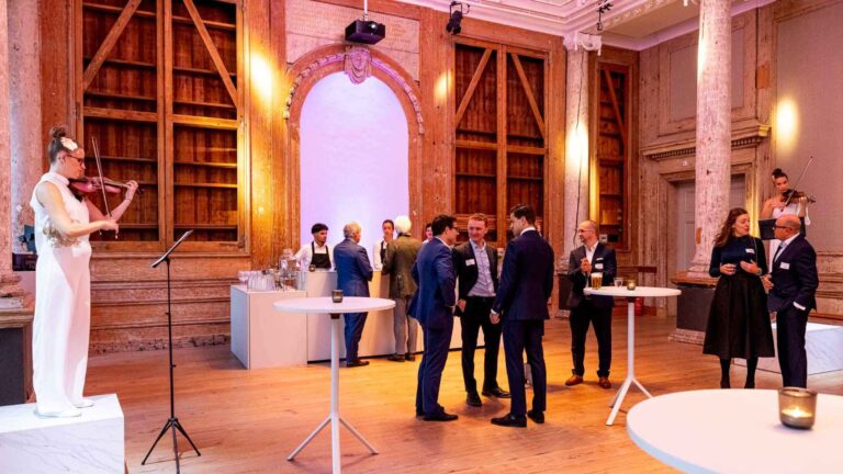 Live violinist performing at a corporate networking reception inside a historic Amsterdam conference venue with exposed wooden beams, classical columns, and guests mingling at standing tables.