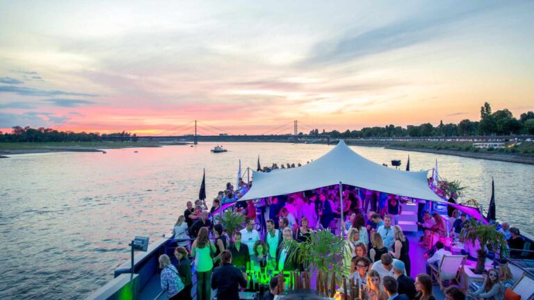 Corporate event guests networking on the upper deck of a river boat venue at sunset, with a white stretch tent canopy and purple ambient lighting reflected across the water.