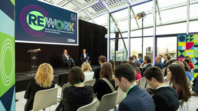 Two speakers in conversation on stage at the Littler REWORK conference in a glass-ceiling venue, with event branding on the main screen and delegates seated in the foreground.