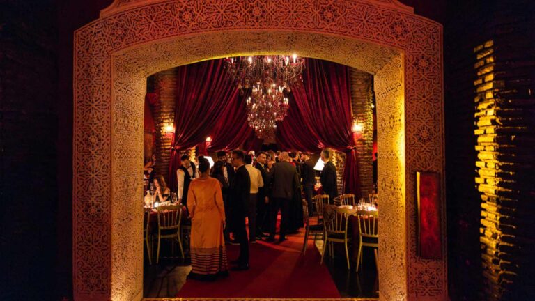 Corporate gala dinner guests viewed through the ornate carved archway at Dar Soukkar in Marrakech, with a chandelier, red velvet drapes, and candlelit tables inside the Moroccan venue.