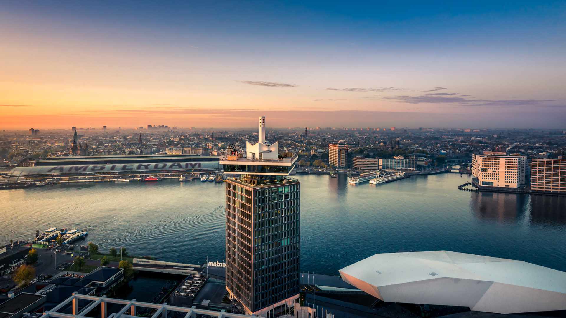 Aerial view of Amsterdam at sunset with the A'DAM Tower and IJ waterway in the foreground, Amsterdam Centraal station across the river, and the Eye Film Museum visible below — a leading corporate event destination in Europe.