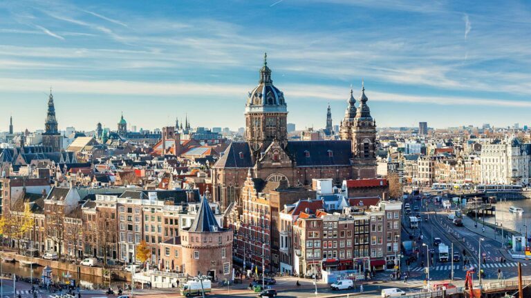 Panoramic view of Amsterdam's historic city centre with the Sint-Nicolaaskerk church and canal house rooftops under a bright blue sky, showcasing one of Europe's top corporate event destinations.