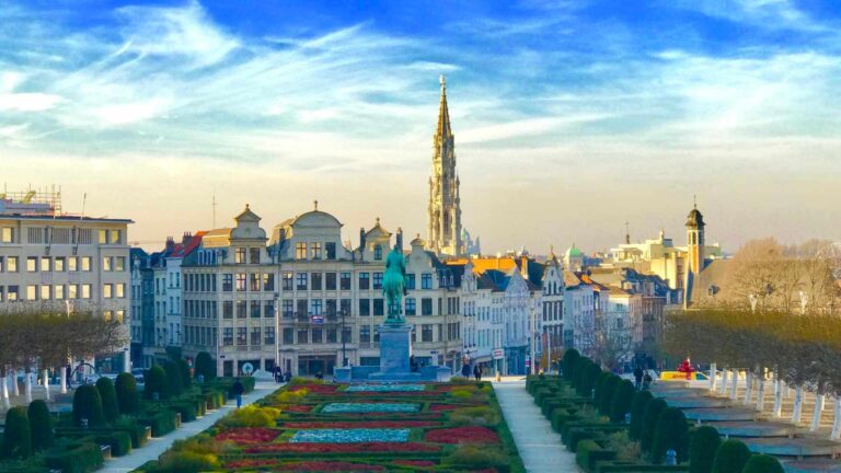 View from Mont des Arts in Brussels with formal colourful gardens, the equestrian statue of King Albert I, and the Brussels Town Hall spire rising above the historic city skyline under a vivid blue sky.
