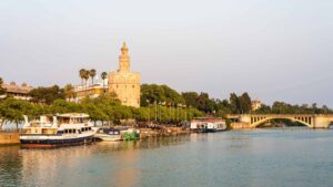 Seville's Torre del Oro watchtower at golden hour, with pleasure boats moored along the Guadalquivir riverside promenade and the Puente de San Telmo bridge arching across the river in the distance.