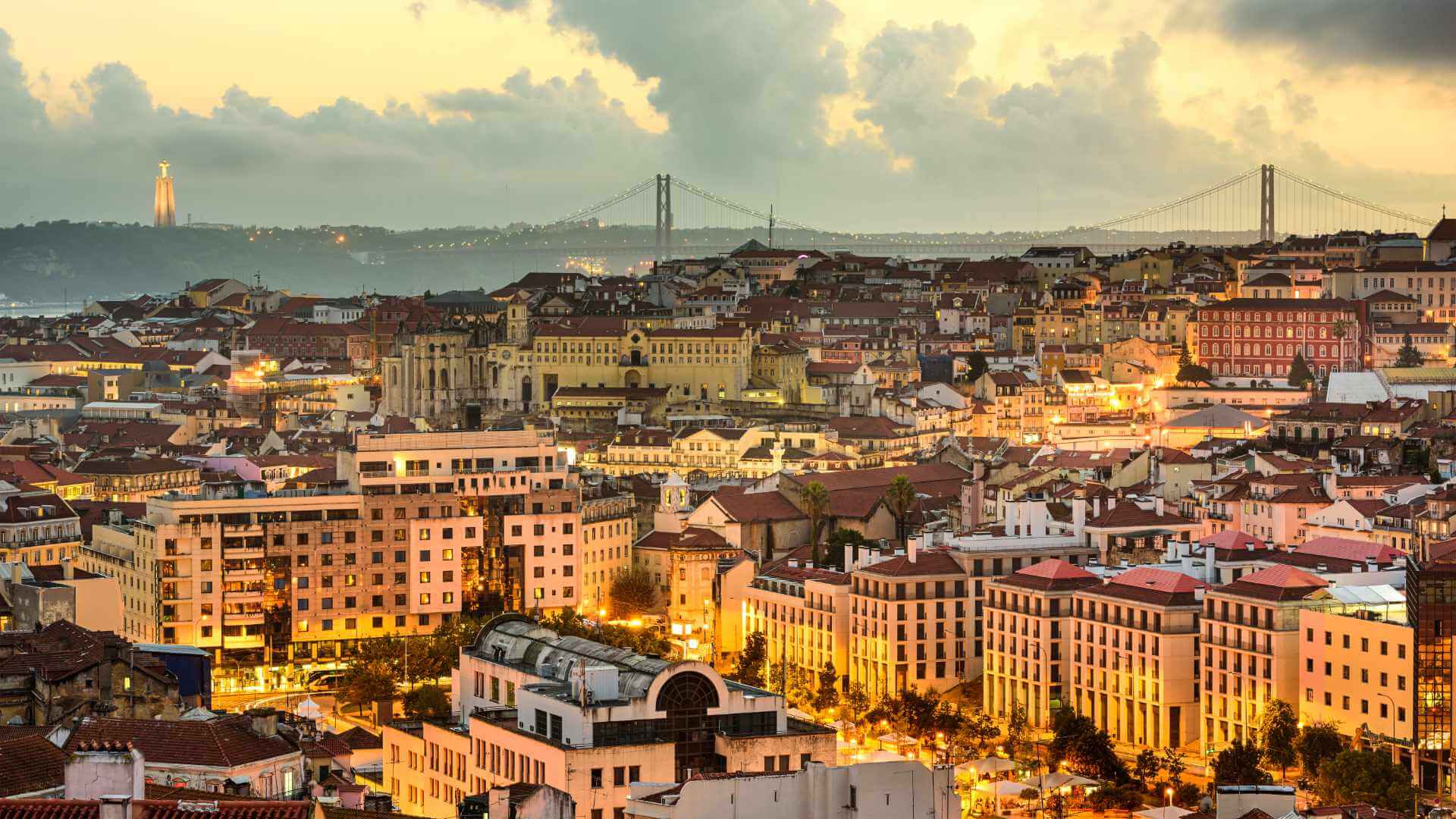Lisbon's illuminated cityscape at night with warm golden lights across the hillside and the 25 de Abril Bridge spanning the Tagus river under a dramatic evening sky.