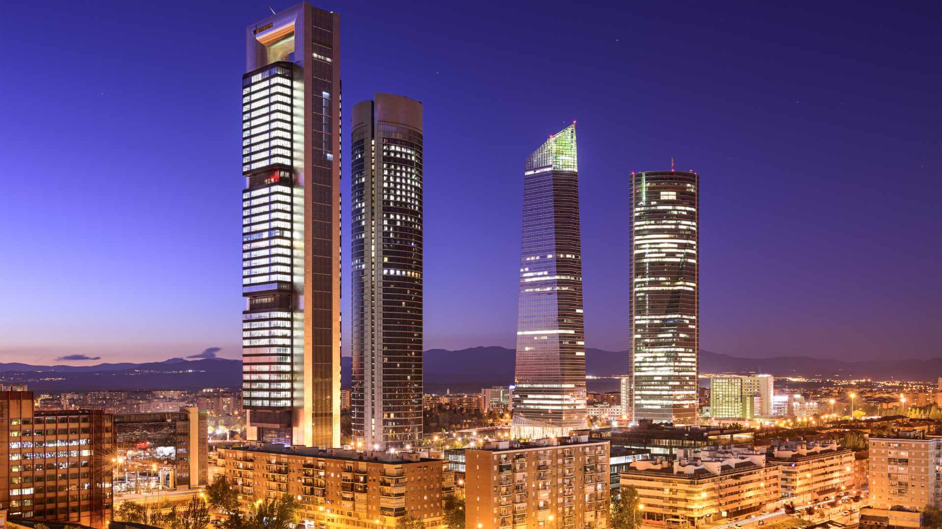 Madrid's Cuatro Torres business district at night, with four iconic skyscrapers glowing against a deep blue twilight sky and the Guadarrama mountains silhouetted in the distance.