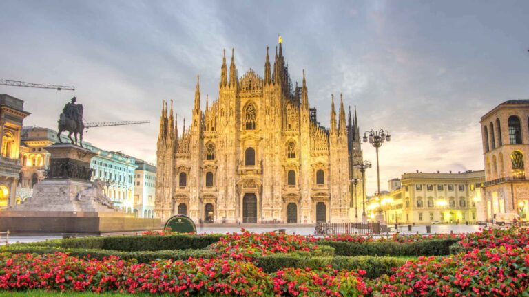 Milan's Duomo di Milano cathedral illuminated at dusk with red flower beds in the foreground, the Victor Emmanuel II equestrian statue to the left, and a dramatic cloudy sky overhead.