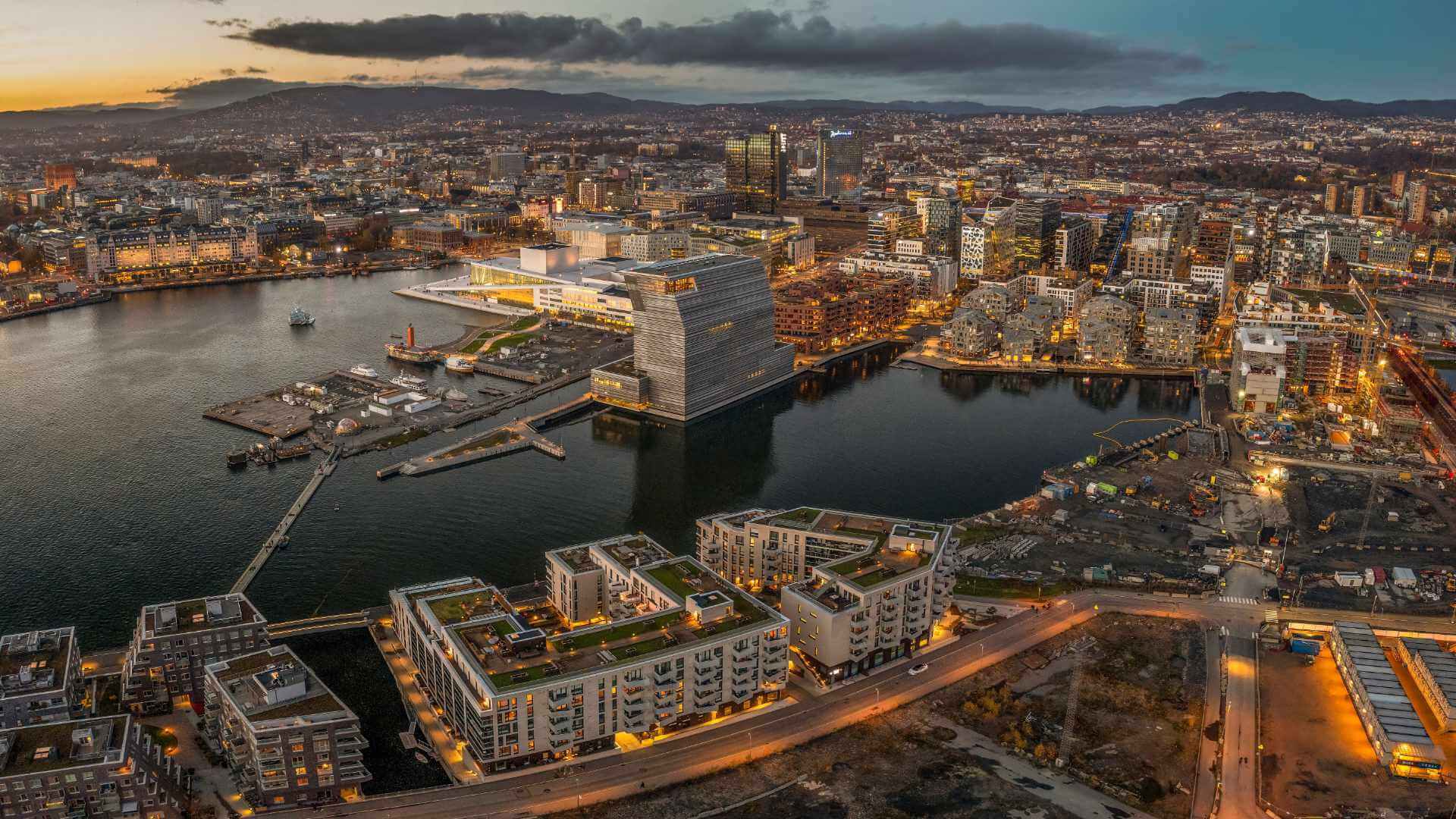 Aerial view of Oslo's Bjorvika waterfront at dusk with the Munch Museum's leaning tower, the Oslo Opera House, and harbour lights reflected across the fjord, with mountains in the background.