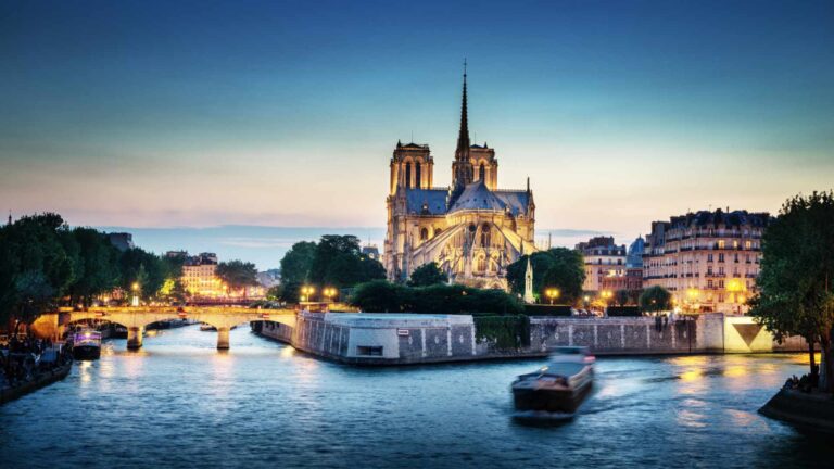 Notre-Dame de Paris cathedral illuminated at night and viewed from across the Seine river, with a barge passing in the foreground and the city's Haussmann buildings glowing under a blue dusk sky.