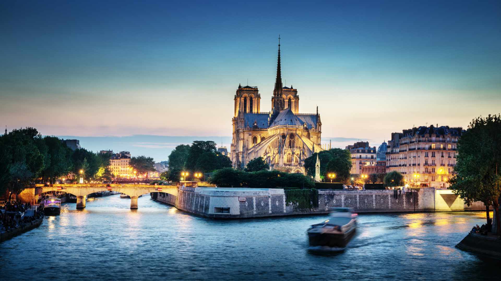 Notre-Dame de Paris cathedral illuminated at night and viewed from across the Seine river, with a barge passing in the foreground and the city's Haussmann buildings glowing under a blue dusk sky.