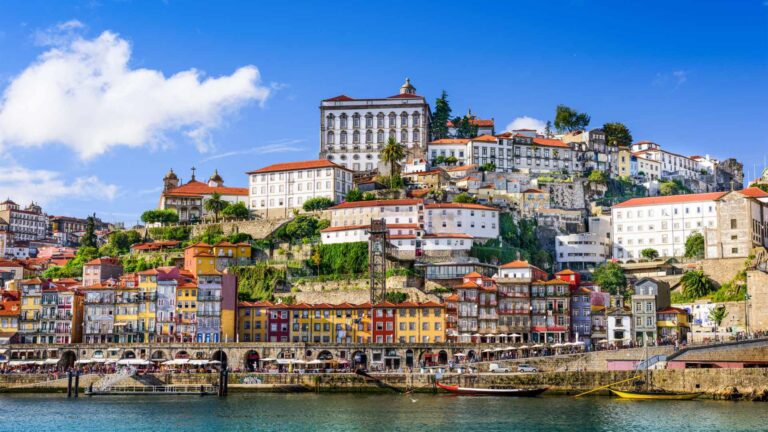 Porto's terraced Ribeira waterfront with colourful historic buildings stacked up the hillside above the Douro river, with traditional Rabelo boats docked at the quay on a sunny day.