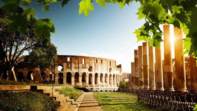 The Colosseum in Rome framed by green maple leaves, with Roman Forum columns visible in the background and sunlight filtering through the trees — an iconic setting for corporate events in Italy.