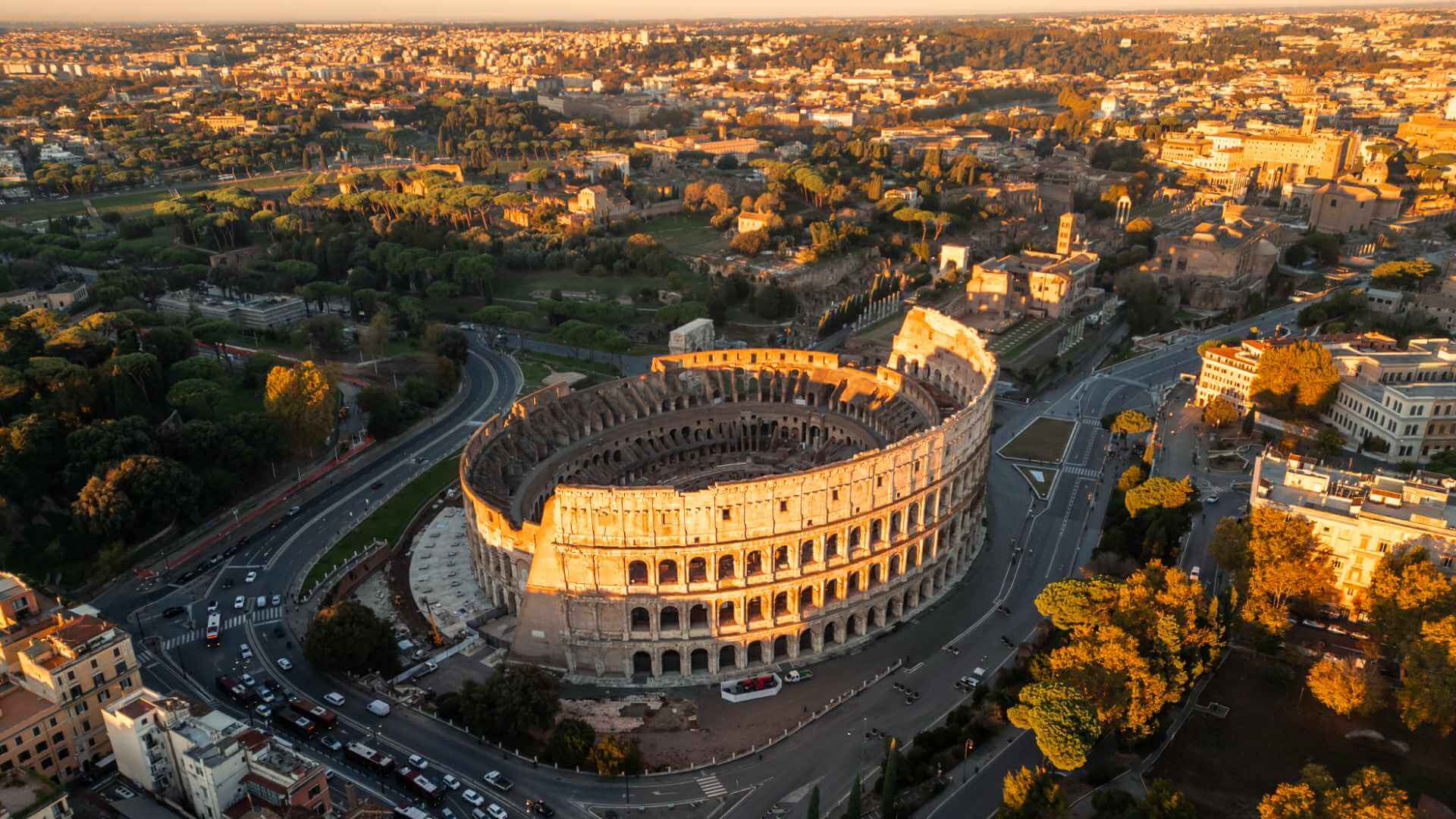 Aerial view of the Colosseum in Rome at golden hour, glowing in early morning light with the Via dei Fori Imperiali and the city's rooftops stretching into the distance.