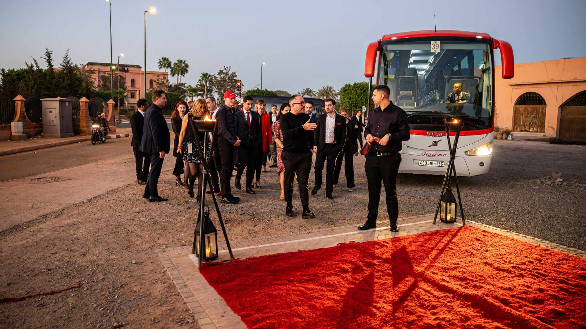 A corporate incentive group arriving at a Moroccan venue in Marrakech at dusk, crossing a red carpet flanked by flame torches as an event manager guides the group from a coach — the coordinated ground transfer and arrival choreography that only a partner with clear ownership of the complete guest journey can deliver.