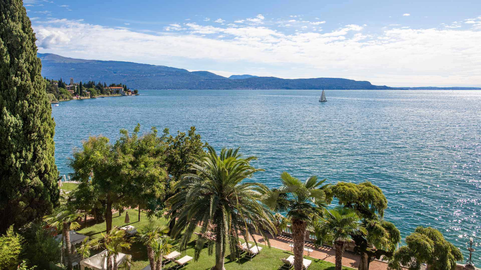 View from a luxury lakeside hotel garden on Lake Garda, Italy, with palm trees, sunbeds and a sailboat on the calm blue water — the kind of destination setting an agency should be able to defend with clear reasoning about why it suits the audience, not just how it photographs.