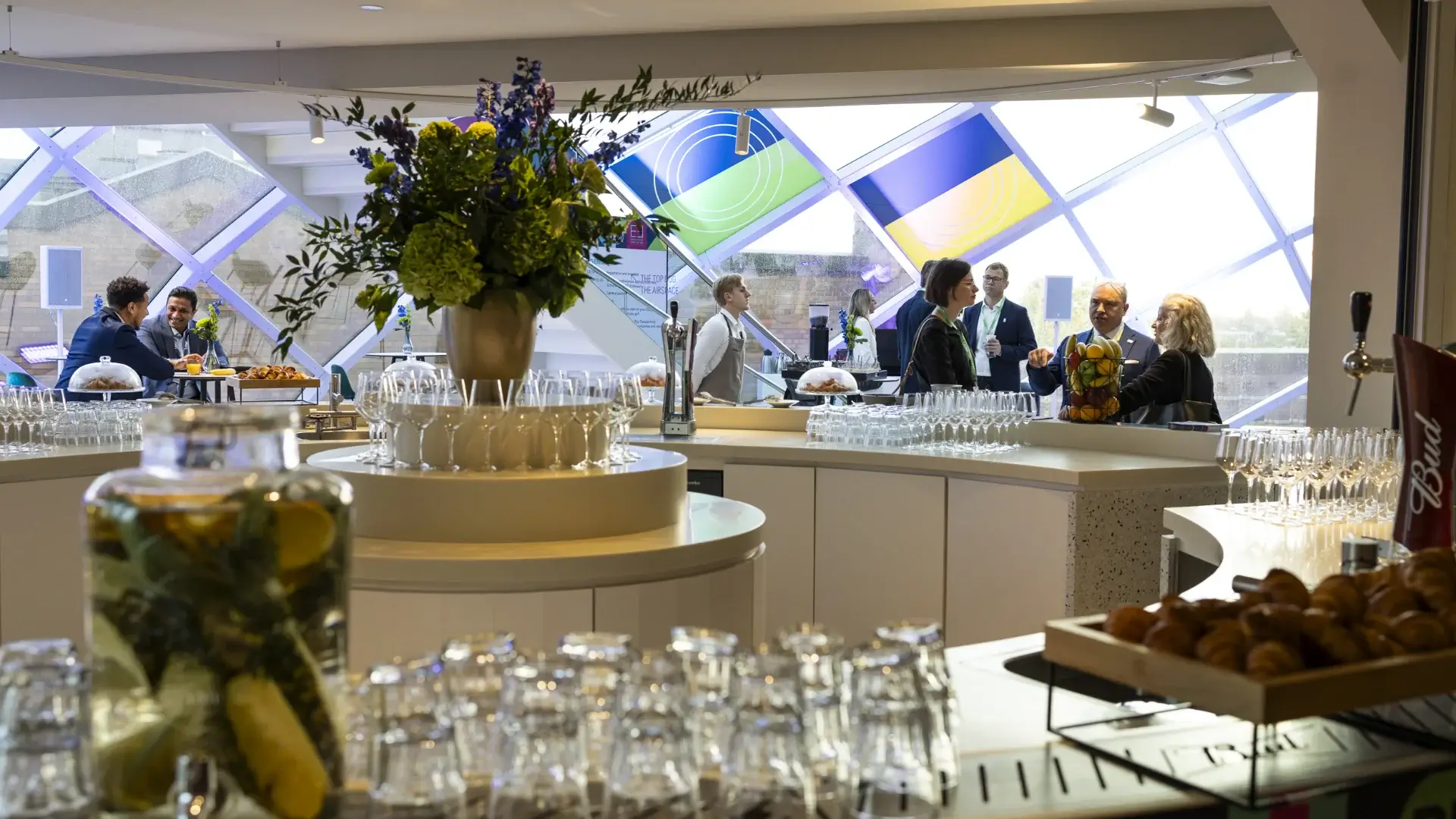 Delegates networking at a corporate event break station — wine glasses, fresh flowers and pastries laid out on a curved bar with a barista — illustrating the catering scope that sits inside a full-service event management fee and varies quietly between proposals that appear to cover the same thing.
