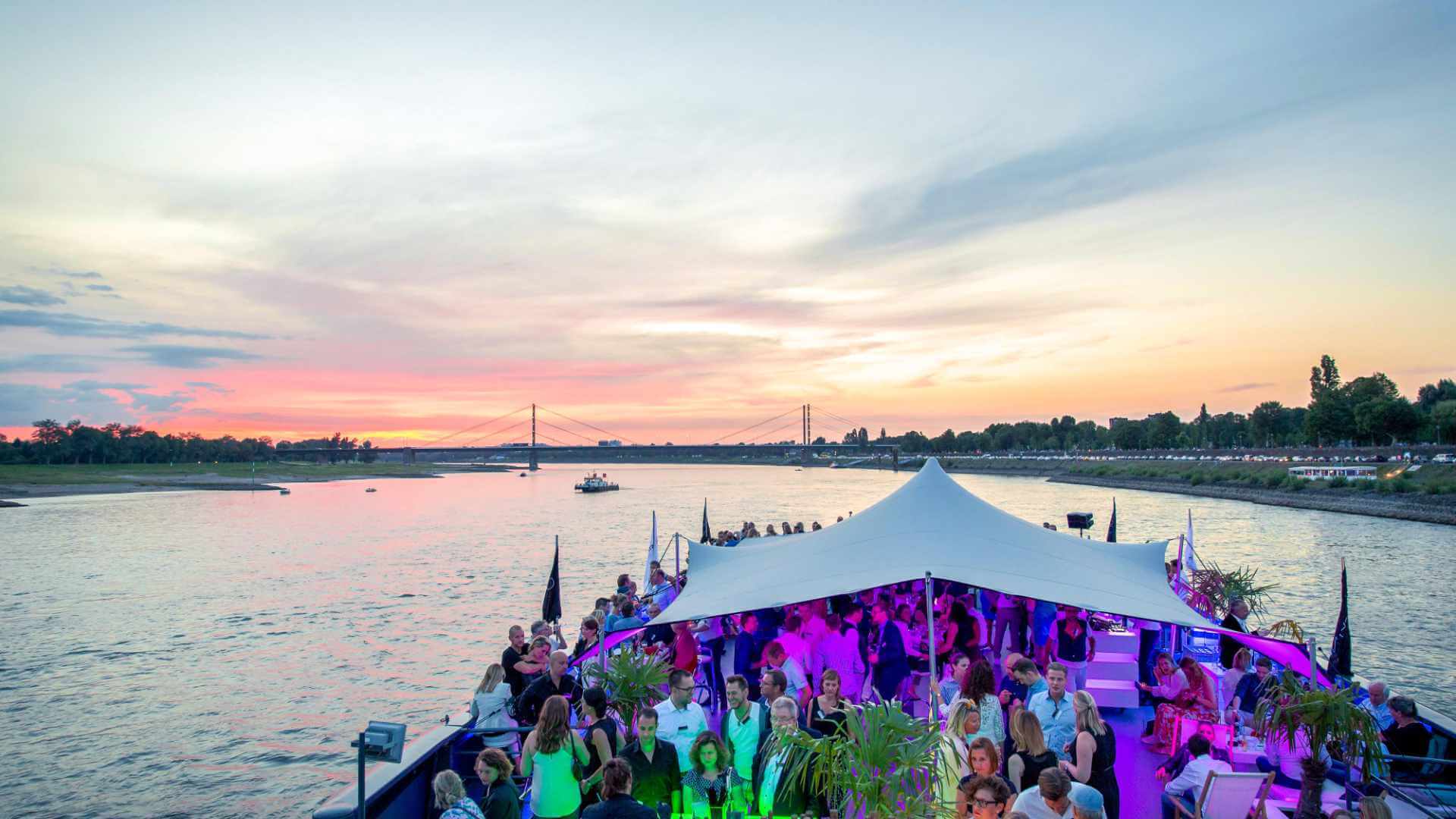 Corporate incentive guests socialising on the lit deck of a private river cruise at sunset, with a stretch tent canopy above and a cable-stayed bridge in the twilight behind — the kind of logistically complex evening movement that tests whether an agency's delivery model can handle transfers, timing and atmosphere simultaneously.