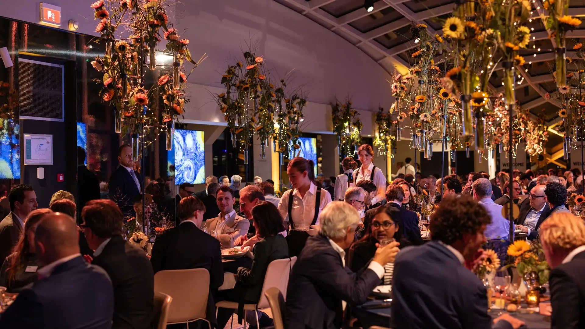 A corporate gala dinner in a Dutch cultural venue, with suspended sunflower floral installations overhead and Van Gogh imagery on the screens — a signature programme moment that makes the case for building a retreat budget around the experiences guests will remember, not a formula applied to every line item equally.