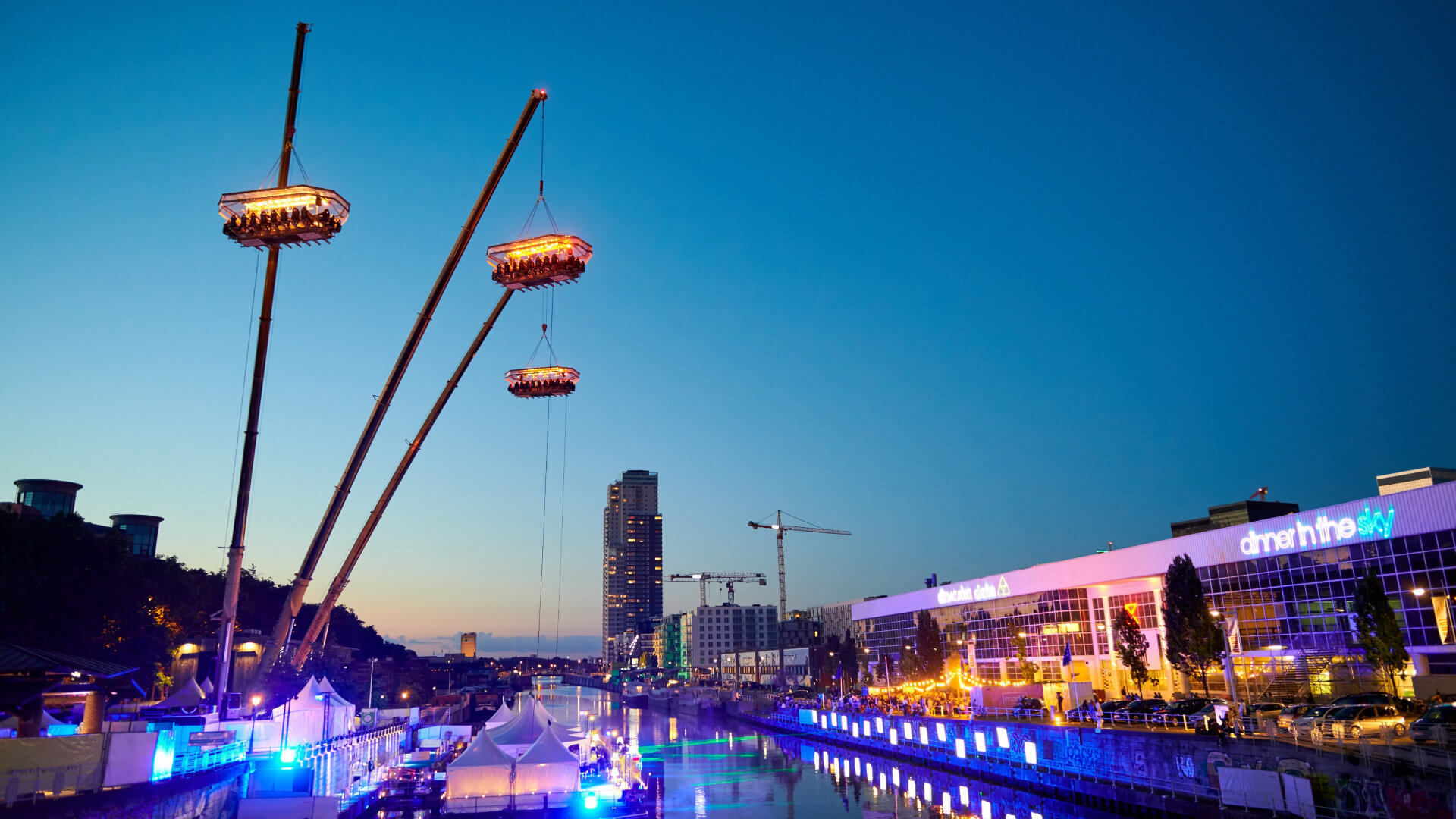 Three illuminated dining tables suspended high above a city at night by cranes in a Dinner in the Sky experience — a high-concept event format that illustrates why incentive travel budgets need transparent pricing, clear permitting and a credible contingency plan before the concept leaves the ground.