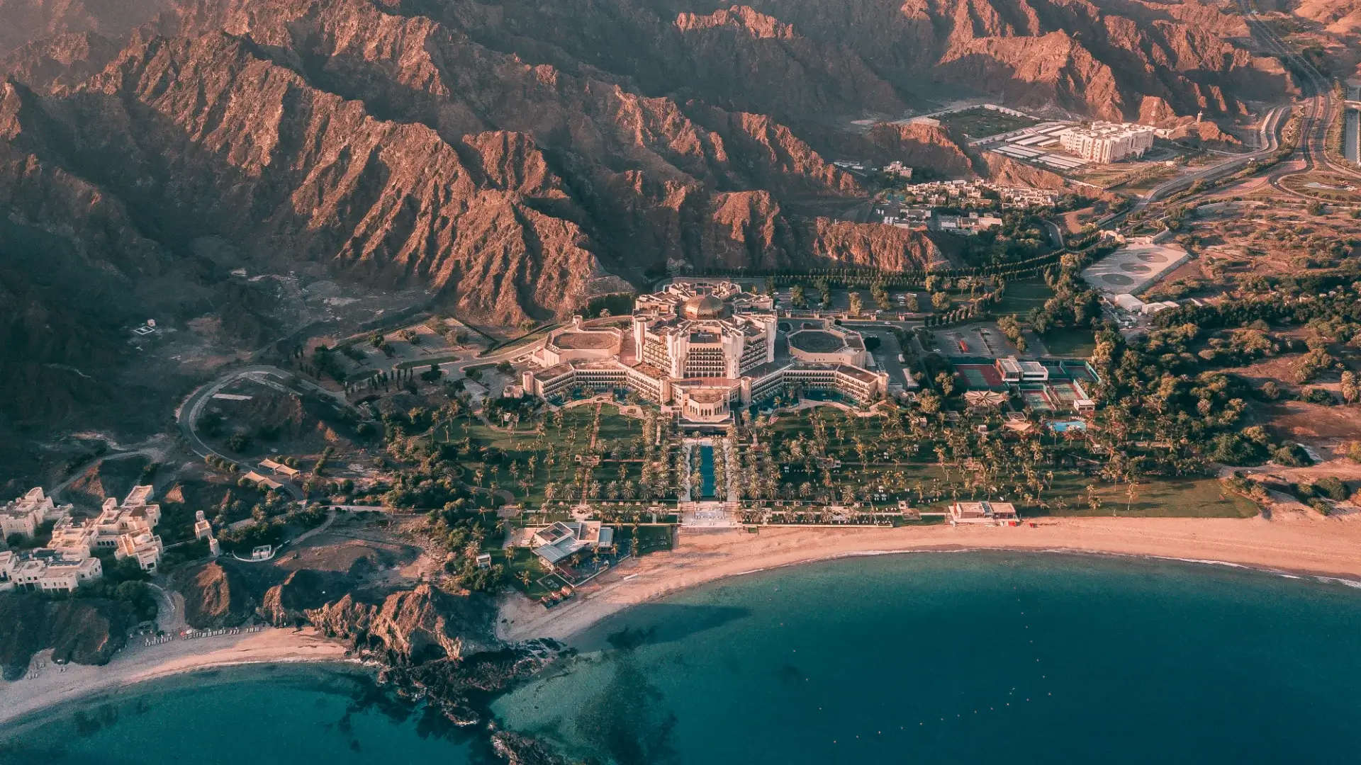 Aerial view of a grand coastal resort in Oman, with a golden-domed main building, palm-lined avenues and a long pool set between the red Hajar mountains and a turquoise bay — a destination where choosing the right property removes the need for transfers and venues, concentrating the budget where guests will actually feel it.
