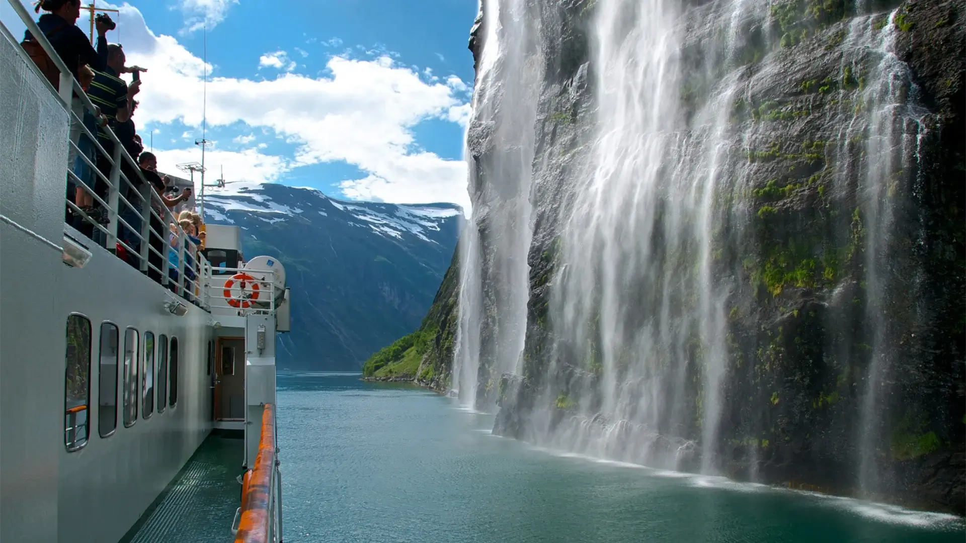 A group on the deck of a cruise vessel in a Norwegian fjord, photographing a waterfall cascading into the turquoise water below, snow-capped peaks above — a destination experience that depends on securing the right vessel, route and season well ahead, and that illustrates why short lead times reduce choice rather than cost.