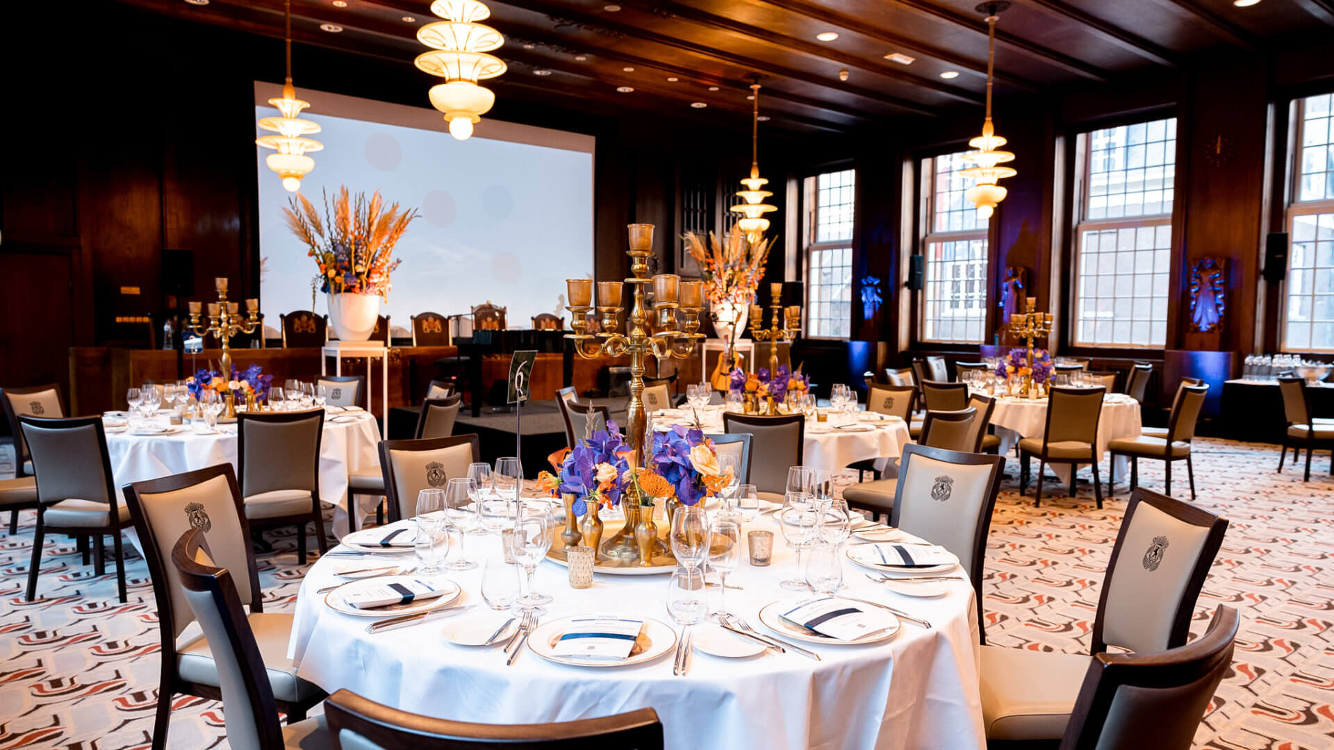 An elegantly dressed corporate dinner venue with round white-linen tables, candelabras, and blue and orange floral centrepieces in a wood-panelled Art Deco dining room — the result when creative direction, venue selection and delivery model are genuinely aligned from the first agency conversation.