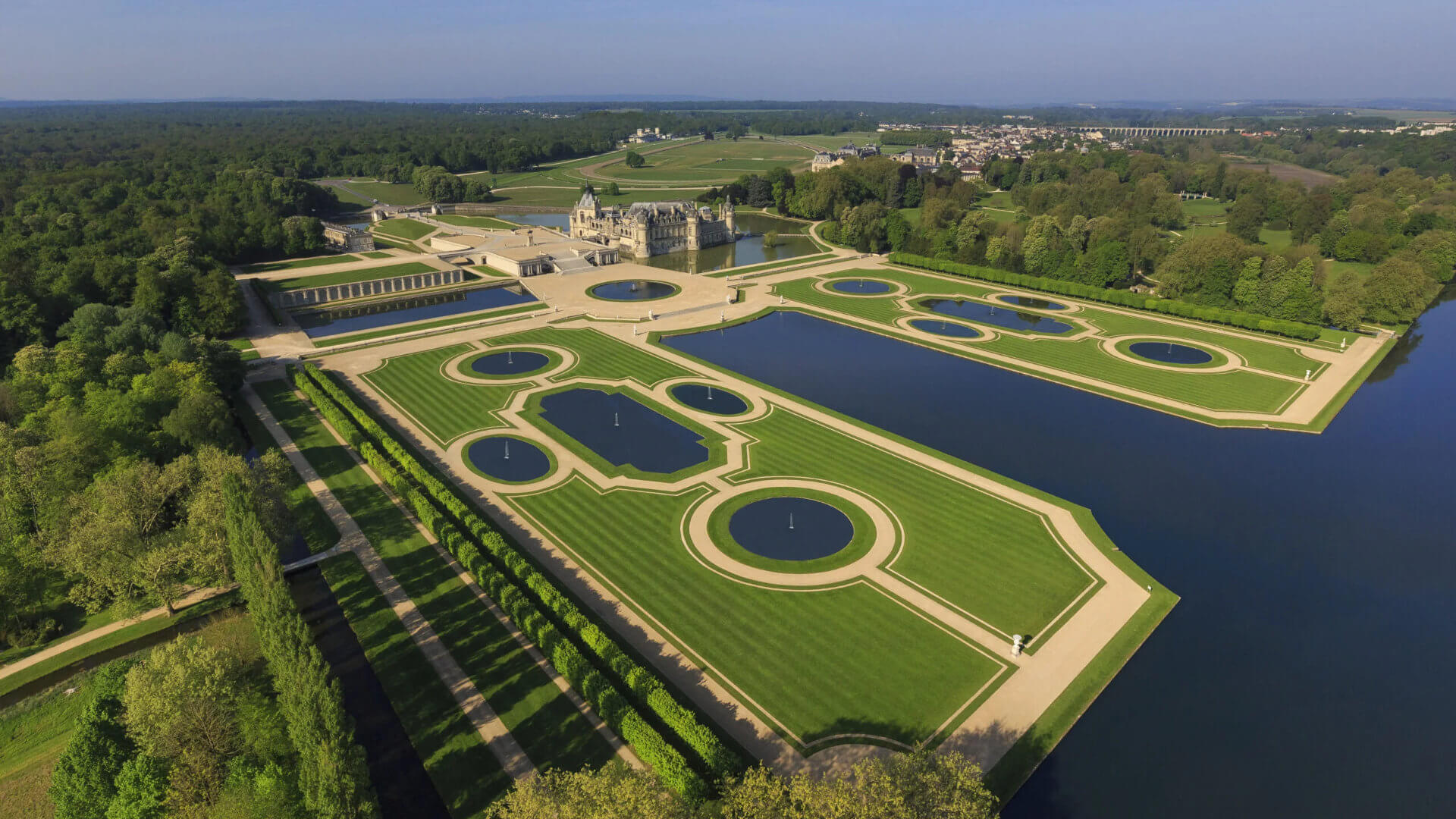 Aerial view of the Château de Chantilly and its formal French gardens — circular fountains, geometric parterres and reflecting pools set within a vast woodland estate — a setting that removes a senior leadership group from the capital and creates the contained environment in which a more focused conversation can take place.