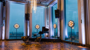 Contemporary Amsterdam event space with floor-to-ceiling windows overlooking the city skyline at dusk, set with a grand piano and decorative globe lights — a contemporary closing venue where an executive dinner can develop into cocktails and music