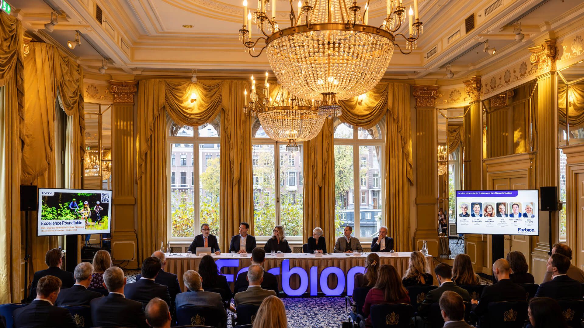 A pharma leadership roundtable in the gilded ballroom of a grand Amsterdam hotel — a panel conversation at a branded table in front of an invited audience — showing the standard of venue and room configuration that a well-planned life-sciences leadership event requires, where every detail of access and atmosphere has been considered in advance.