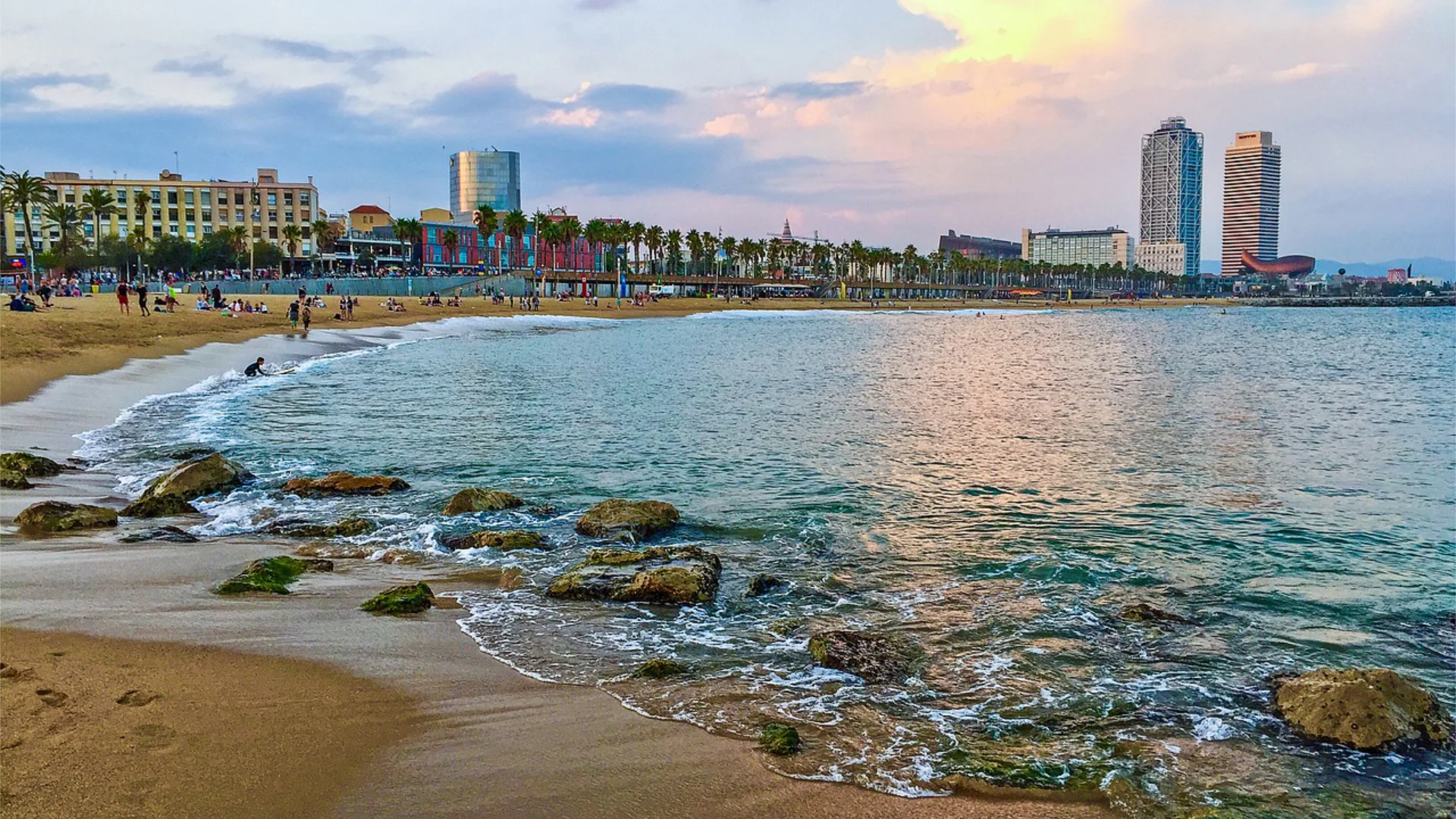 Barceloneta beach at sunset, with the Hotel Arts and Torre Mapfre towers rising above the palm-lined promenade and the Mediterranean stretching to the horizon — the waterfront that connects El Tribut, Marina Port Vell and SALT into a single coherent Barcelona guest journey when each venue has one clear role in the programme.
