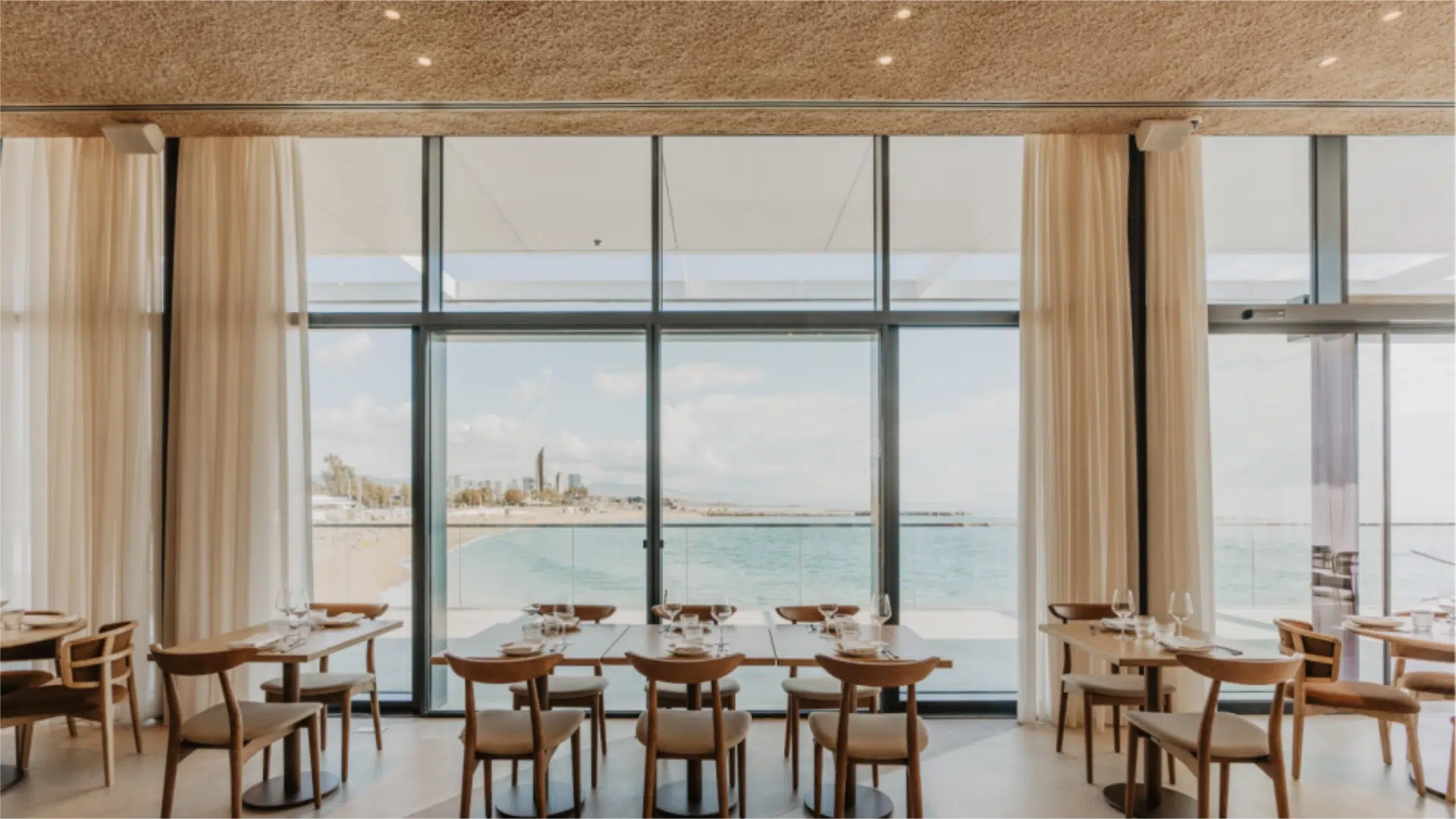 The dining room at El Tribut in Port Olímpic, Barcelona — cork ceiling, linen curtains and wooden tables set with wine glasses, floor-to-ceiling windows framing a direct Mediterranean sea view — an interior that delivers a clear sense of place without requiring a programmed cultural layer on top of it.