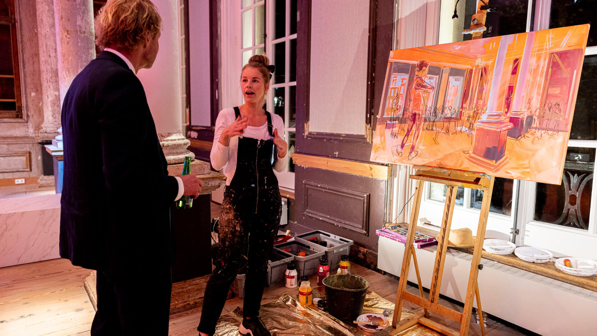 Live artist in paint-splattered dungarees discussing her work-in-progress canvas with a corporate guest in an Amsterdam canal-house interior — the kind of private cultural access that gives a CEO programme genuine local depth beyond familiar sightseeing