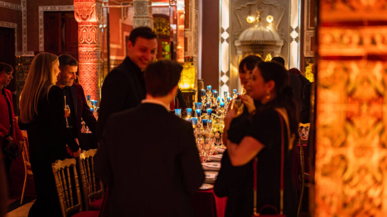 Corporate incentive guests gathering at a candlelit gala dinner table in a richly decorated Marrakech riad, with ornate zellige columns and blue candle glasses — the private city dining experience where structure disappears and conversation takes over