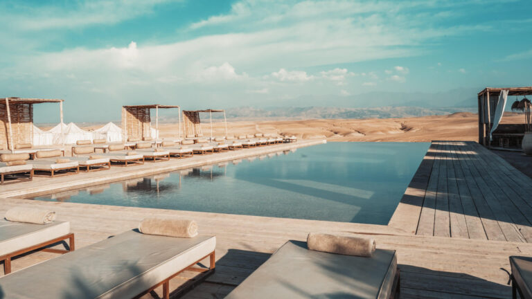 Luxury infinity pool at an Agafay desert camp with sun loungers, reed shade canopies and the Atlas Mountains in the background — the change of landscape and perspective that makes a journey beyond Marrakech worth the access planning it requires