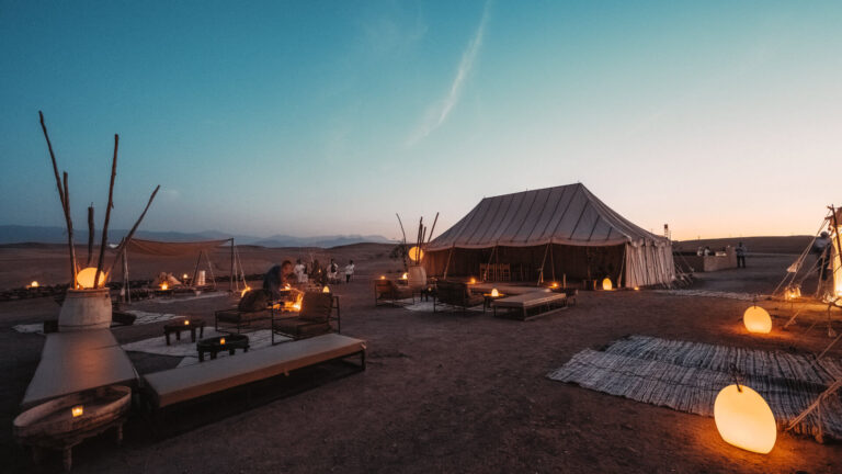 Aerial view of a large Berber-style incentive event camp being prepared at sunset near Marrakech, with white marquee tents, red carpet and production vehicles — the operational scale behind what guests experience as a seamless desert evening