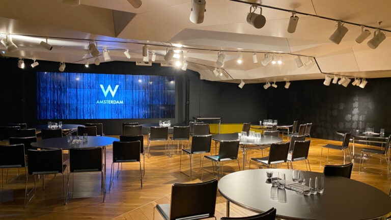 A corporate event space set in cluster format at W Amsterdam, with round black tables, track lighting, herringbone parquet and the hotel's LED video wall — an example of how venue briefing and room layout translate a programme's intended atmosphere into the physical space.