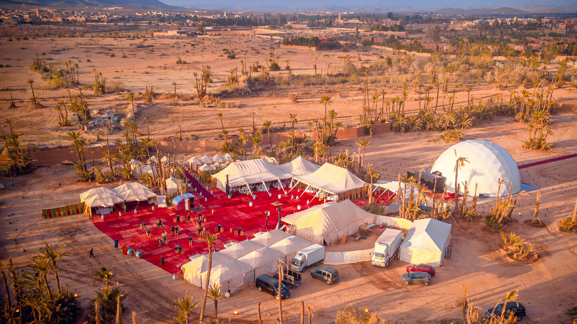 Aerial view of a large corporate event camp near Marrakech at dusk, with traditional white tents, a geodesic dome and palm trees — every journey beyond the city should add something the Medina cannot provide and must justify the movement it creates