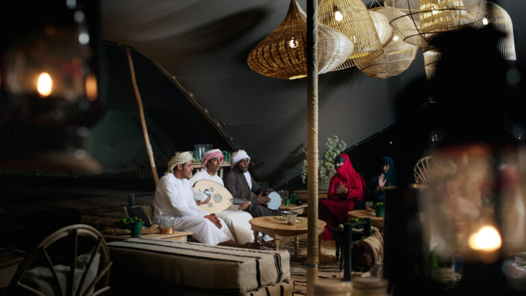 Traditional Omani musicians performing with an oud and drum beneath a Bedouin-style tent at night, with guests in traditional dress listening nearby — an authentic local cultural encounter that was genuinely possible because an agency tested the concept against the destination's real conditions before committing it to the itinerary.