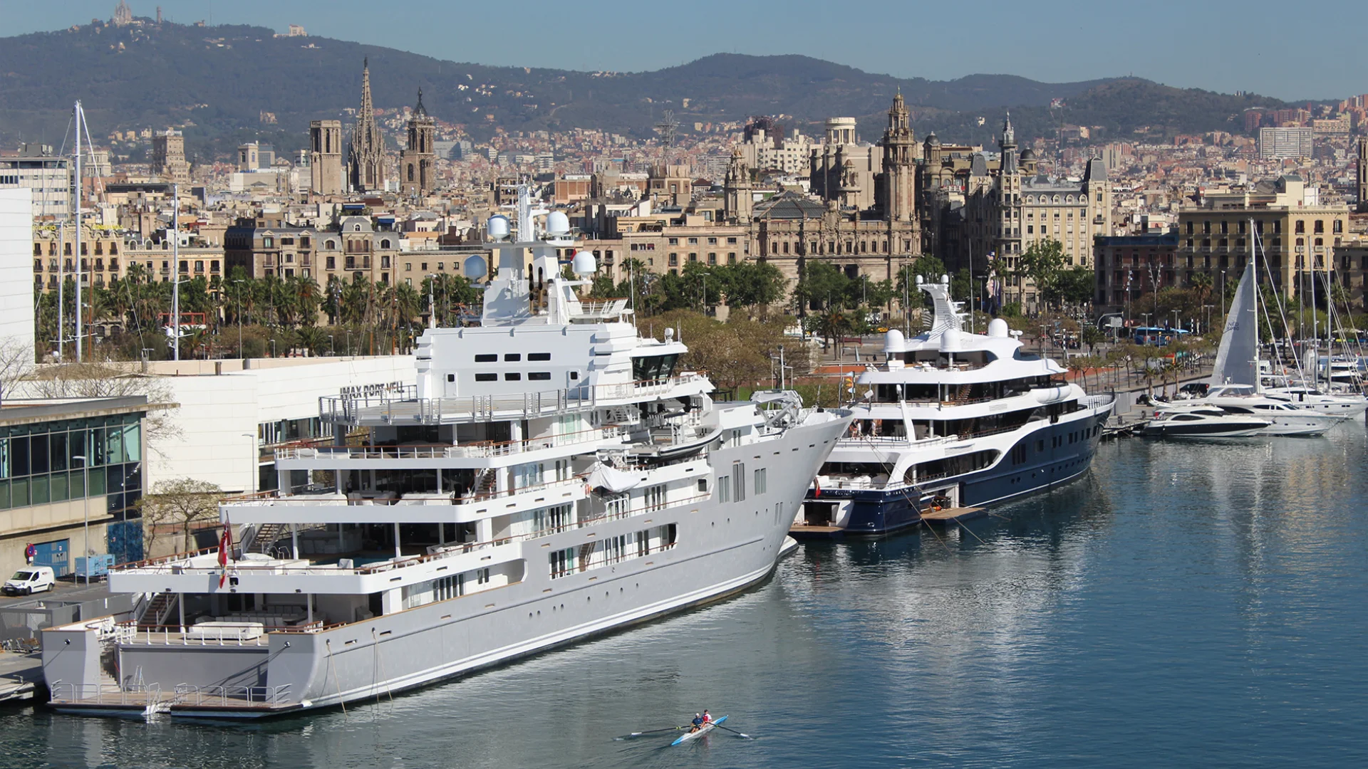 Superyachts moored at Marina Port Vell in Barcelona, with the Gothic Quarter and Montjuïc rising above the city skyline behind — a marina whose visual environment sets an internationally recognisable tone for a principal client event before any production element arrives.