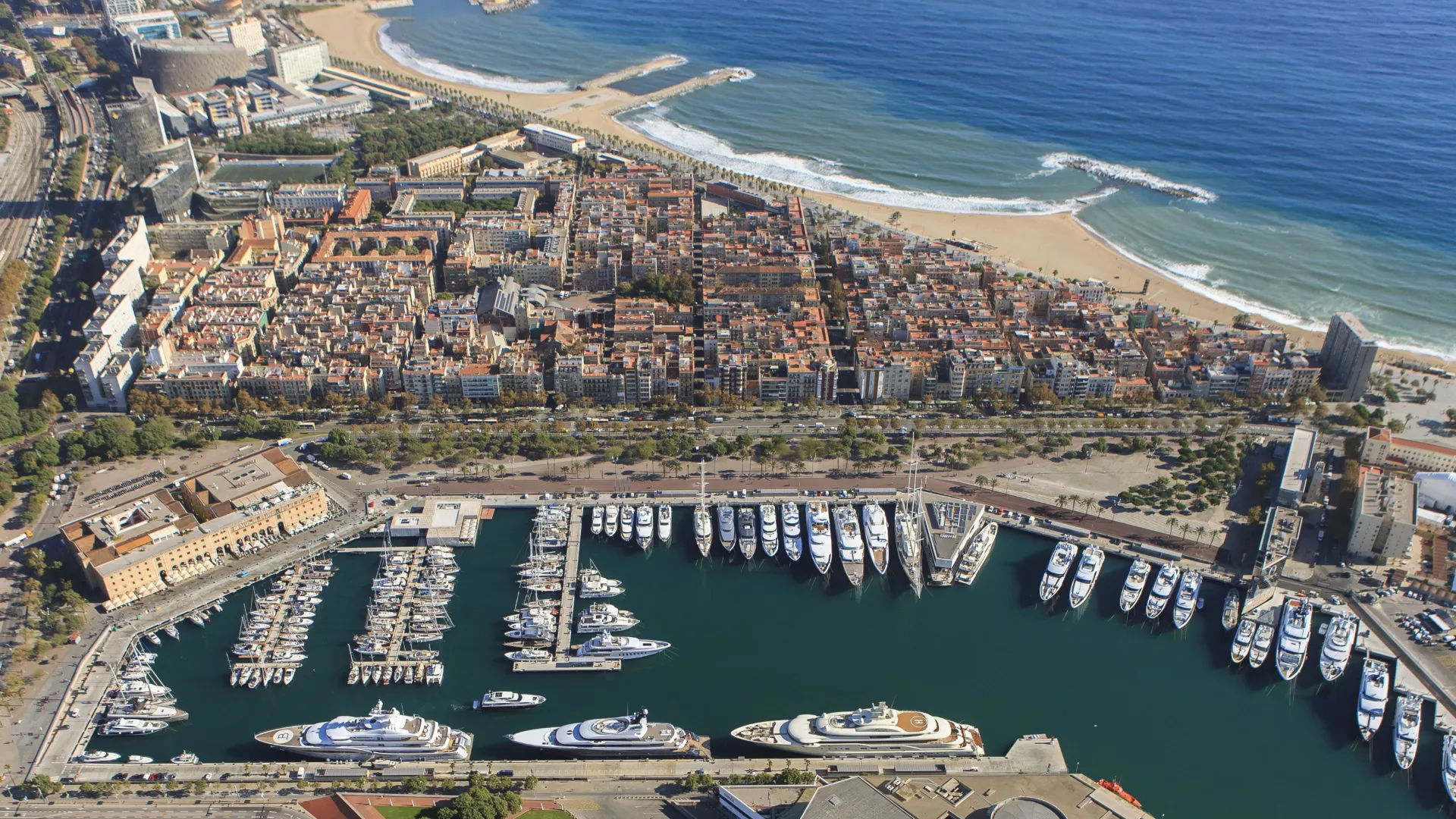 Aerial view of Marina Port Vell and the Barcelona waterfront — the superyacht marina between the Barceloneta neighbourhood and the Mediterranean, with the coastline stretching toward Port Olímpic and W Barcelona — showing the compact geography that makes a three-venue waterfront programme operationally viable in a single evening.