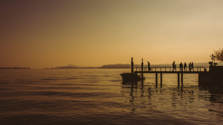 Silhouettes of guests on a wooden Italian lake jetty at sunset, with a boat moored alongside and distant mountains reflected in the amber water — the unstructured pause that a well-paced incentive travel programme builds into the itinerary, and that guests remember long after the scheduled activities.