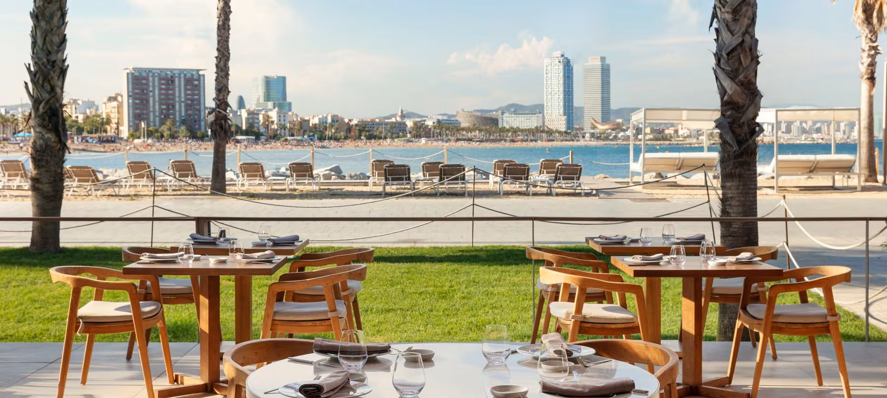 The beachside terrace at SALT at W Barcelona — wooden chairs and set tables on a coastal lawn, with the Hotel Arts, Torre Mapfre and Frank Gehry's fish sculpture across the bay — a venue that can bring informality to the programme's final chapter while retaining the service infrastructure of an established international hotel.