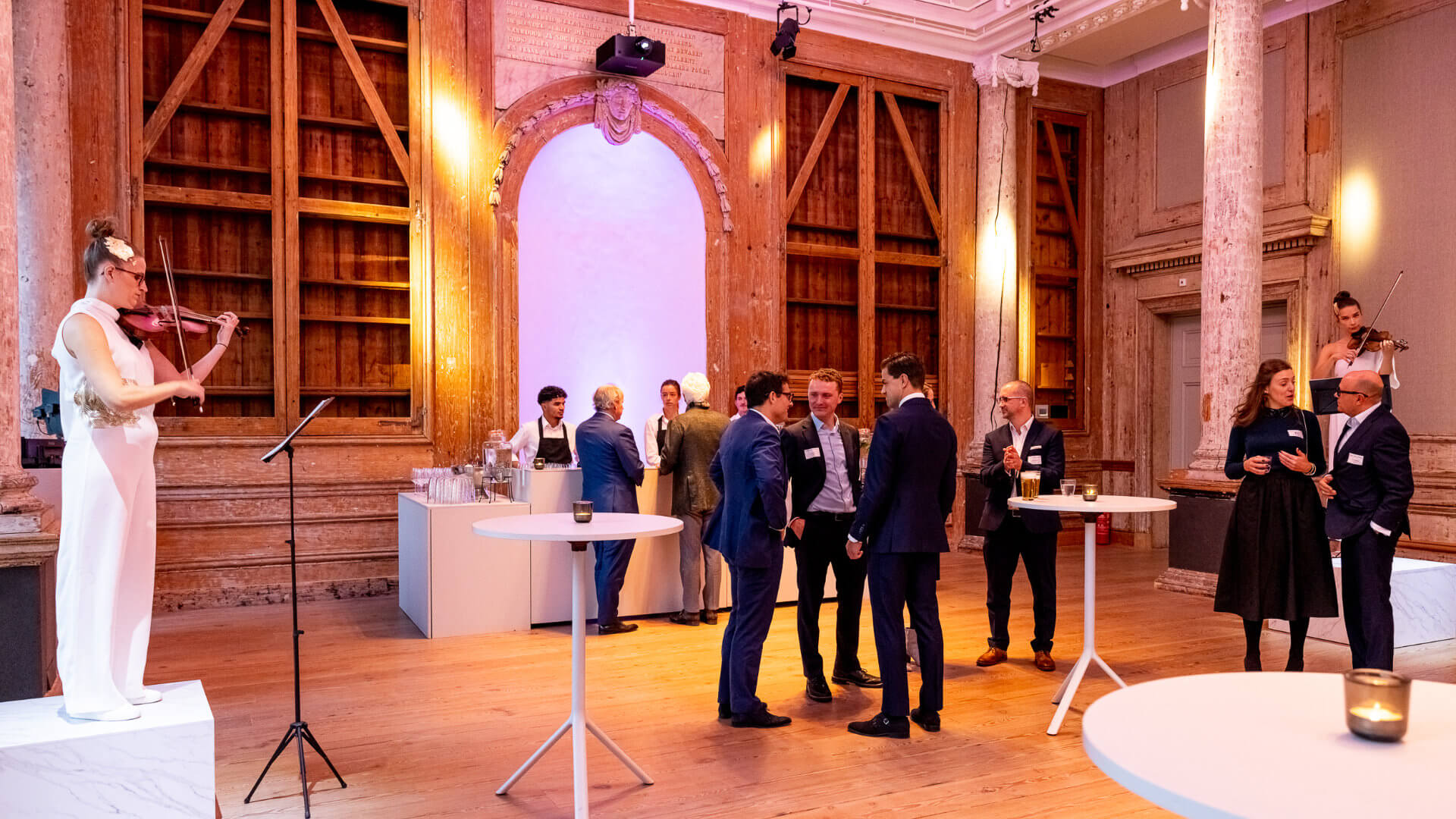 Executive welcome reception in a landmark Amsterdam hall with two violinists performing and guests networking at cocktail tables — the opening moment of a CEO programme designed to settle the group before the leadership agenda begins
