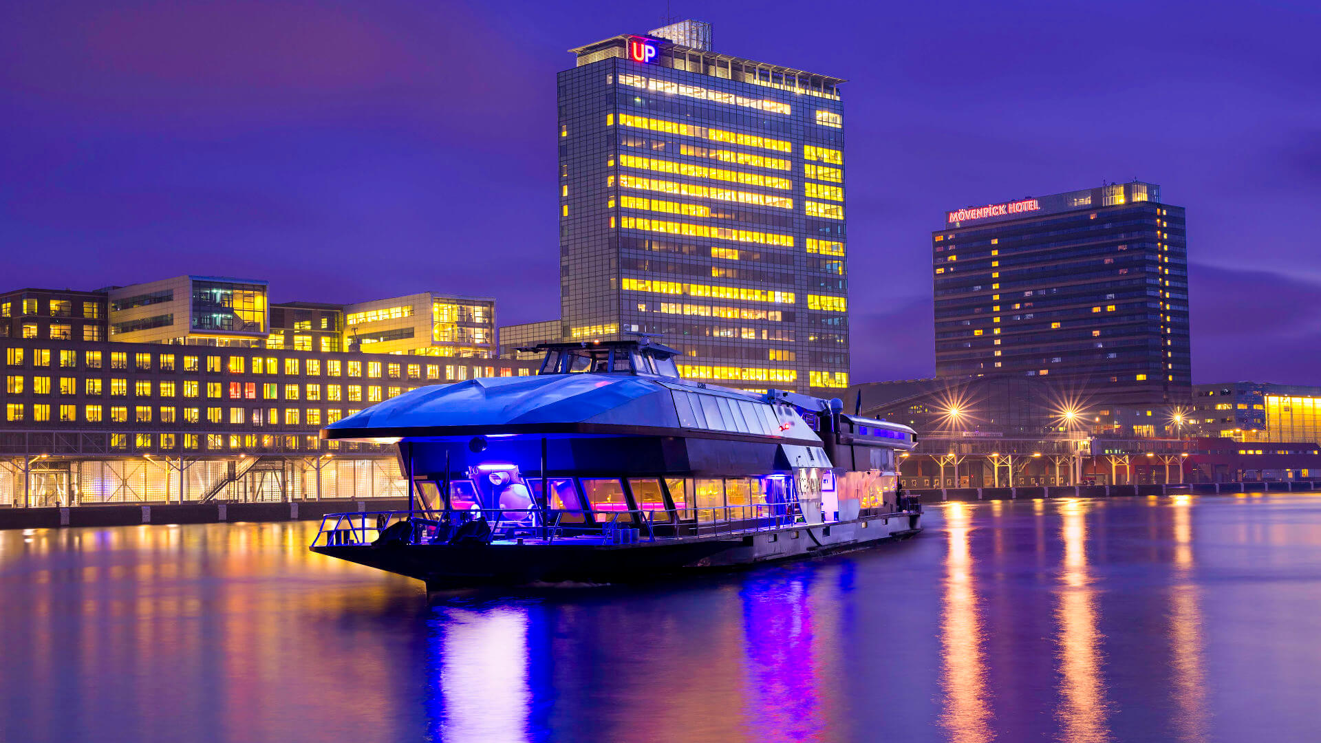 Private executive salon boat moored on Amsterdam's IJ waterway at night with the North Amsterdam skyline behind — water transport that provides a contained, private environment between programme venues rather than a narrated sightseeing break
