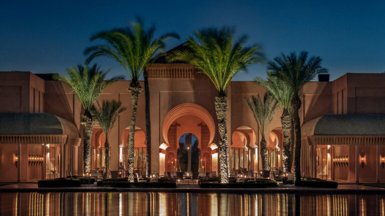 Grand Marrakech luxury hotel illuminated at night with Moorish arches, palm trees and a reflecting pool — the base whose location shapes the entire transport and logistics plan for an incentive group