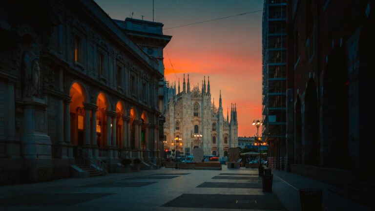 The Milan Duomo framed by a street approach at sunset, its white marble spires lit against a deep orange sky and the Palazzo della Ragione arcades glowing amber to the left — the concentrated character of a city where architectural history and contemporary design occupy the same compact centre.