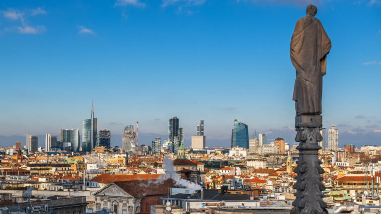The modern Milan skyline viewed from the Duomo rooftop — a Gothic stone statue in the foreground with the UniCredit Tower and Porta Nuova business district rising beyond the terracotta rooftops — illustrating why Milan carries genuine business credibility without feeling like a conventional conference city.