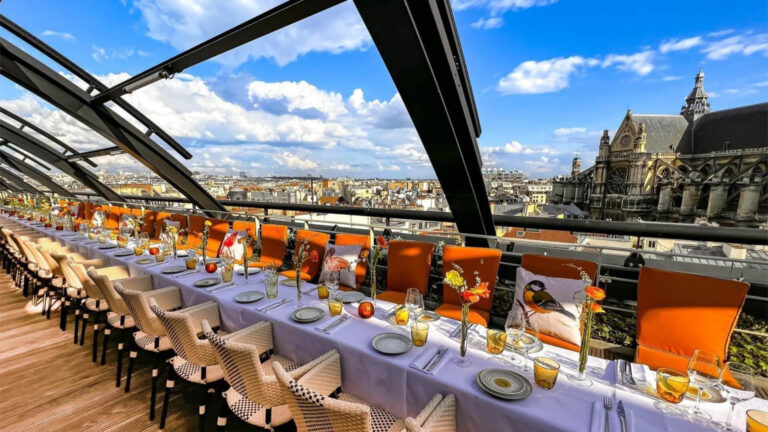 A private rooftop dinner above the Paris skyline — a long table with orange chairs and bird-print cushions under a retractable glass roof, with the Church of Saint-Eustache and Haussmannian rooftops stretching to the horizon — the kind of one-of-a-kind setting that gives a CEO programme a reason to visit Paris that goes beyond a five-star hotel reservation.