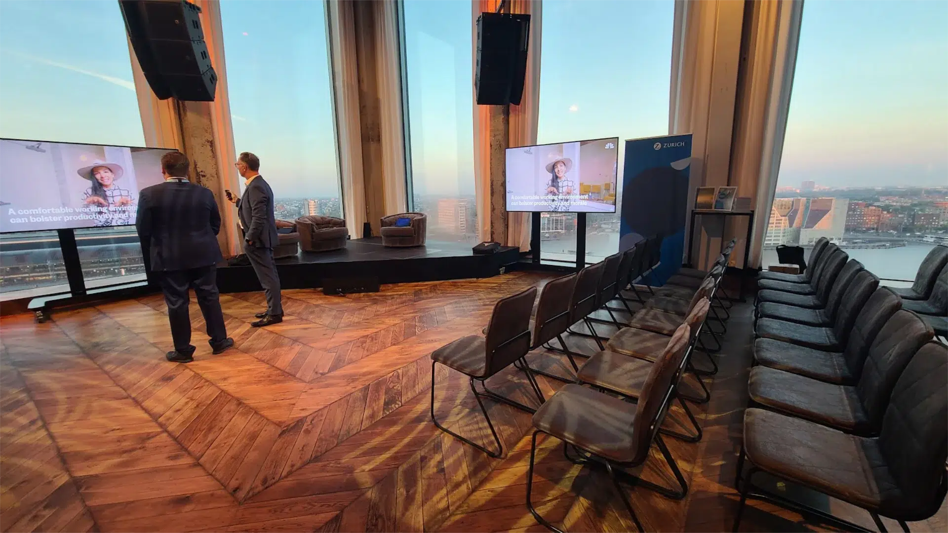 High-rise event venue in Amsterdam with floor-to-ceiling city views, theatre-style seating and presentation screens, illustrating why an evening session that adds external guests to the core group needs its own separate logistics plan
